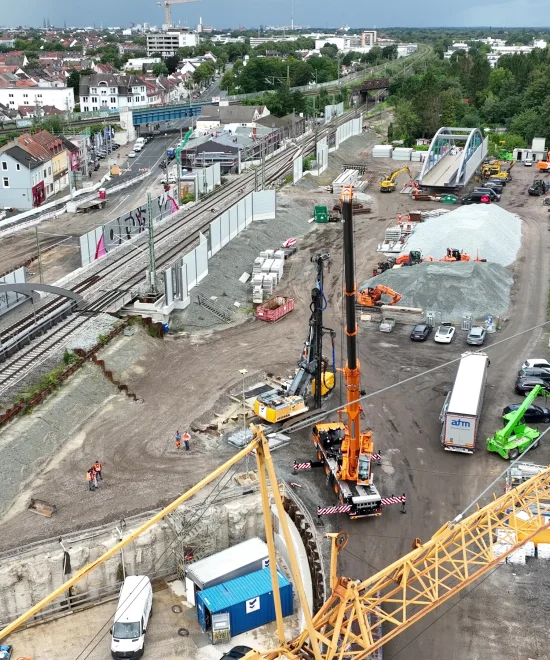 Luftaufnahme einer Bahnbaustelle mit Brückenbau, Gleisarbeiten und Kränen in einem Stadtgebiet – zeigt die komplexe Infrastrukturplanung und Bauüberwachung der ai Consult GmbH.